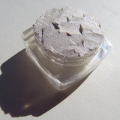 Glittery eyeshadow in a clear plastic jar, silver-white color, textured surface, angled shadow