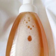 Foaming soap bottle with white pump, amber liquid, and white foam bubbles on white fabric