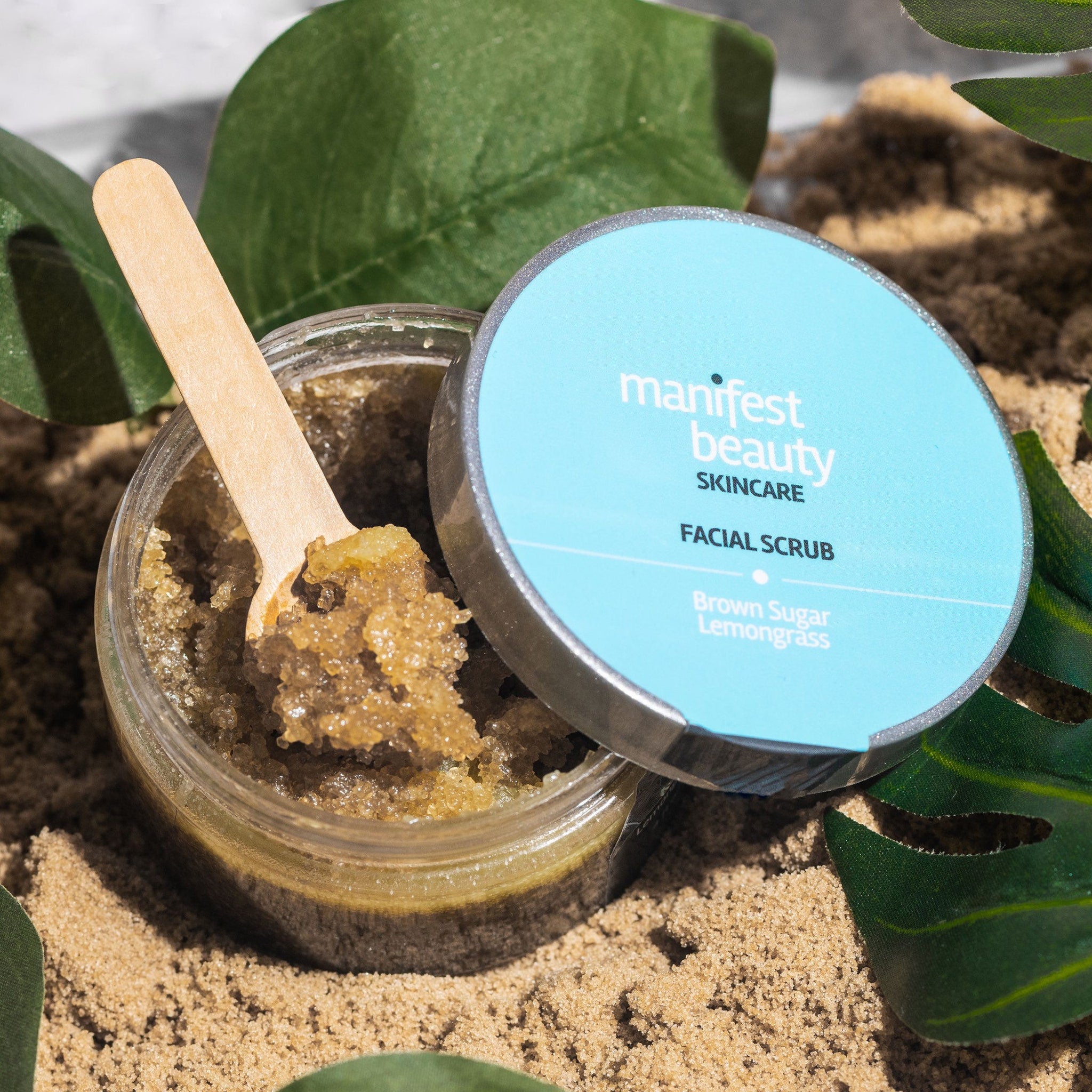 Facial scrub in clear jar with brown granules and wooden spoon, Manifest Beauty brand