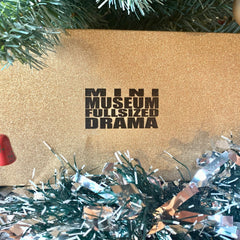 Gold glittery box with black text "MINI MUSEUM FULLSIZED DRAMA" on a Christmas background