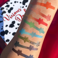 Eyeshadow swatches in various colors, including glitter and matte finishes, applied to arm with "Vicious D" text in background