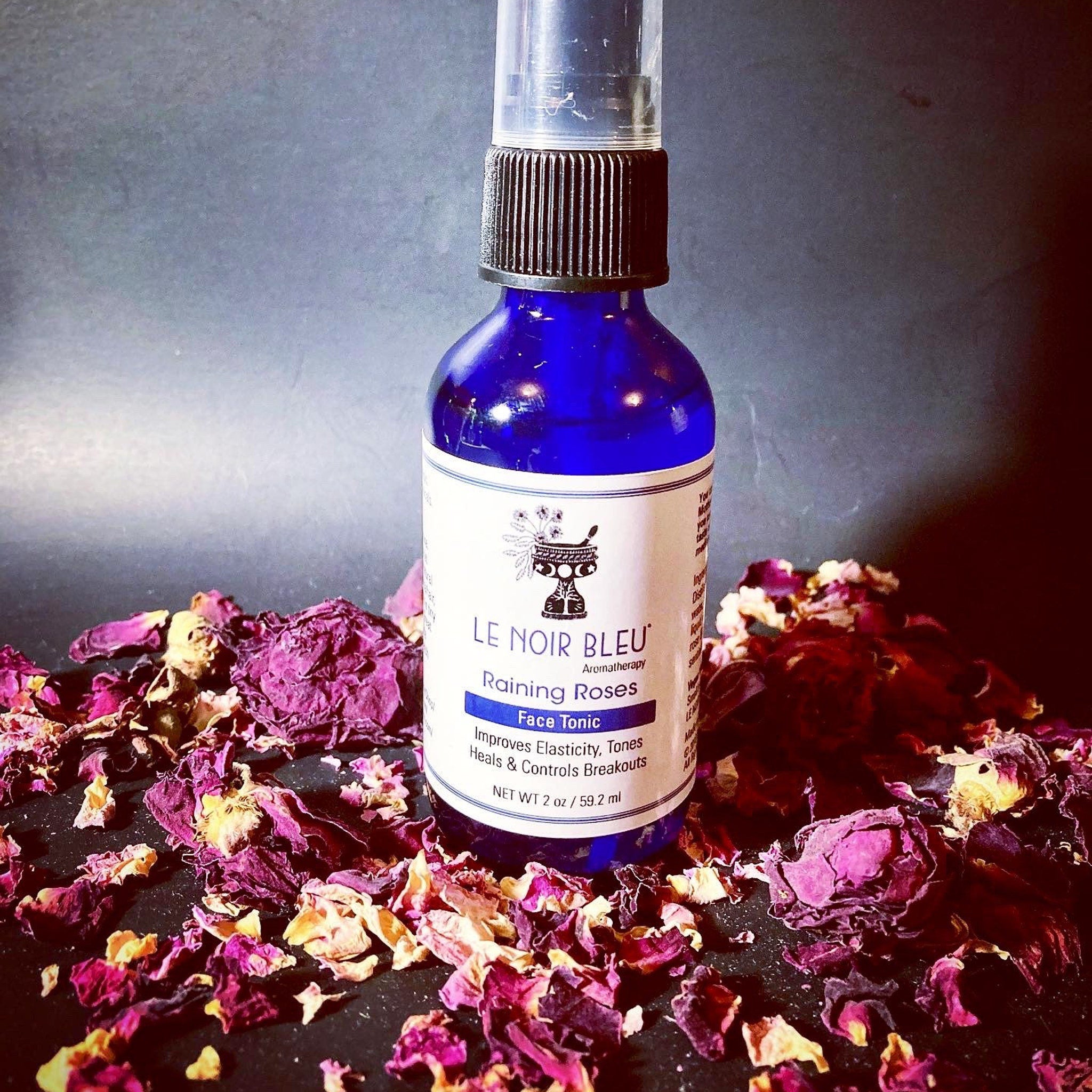 Toner bottle on black surface, blue glass bottle with black cap, surrounded by rose petals, Le Noir Bleu label visible