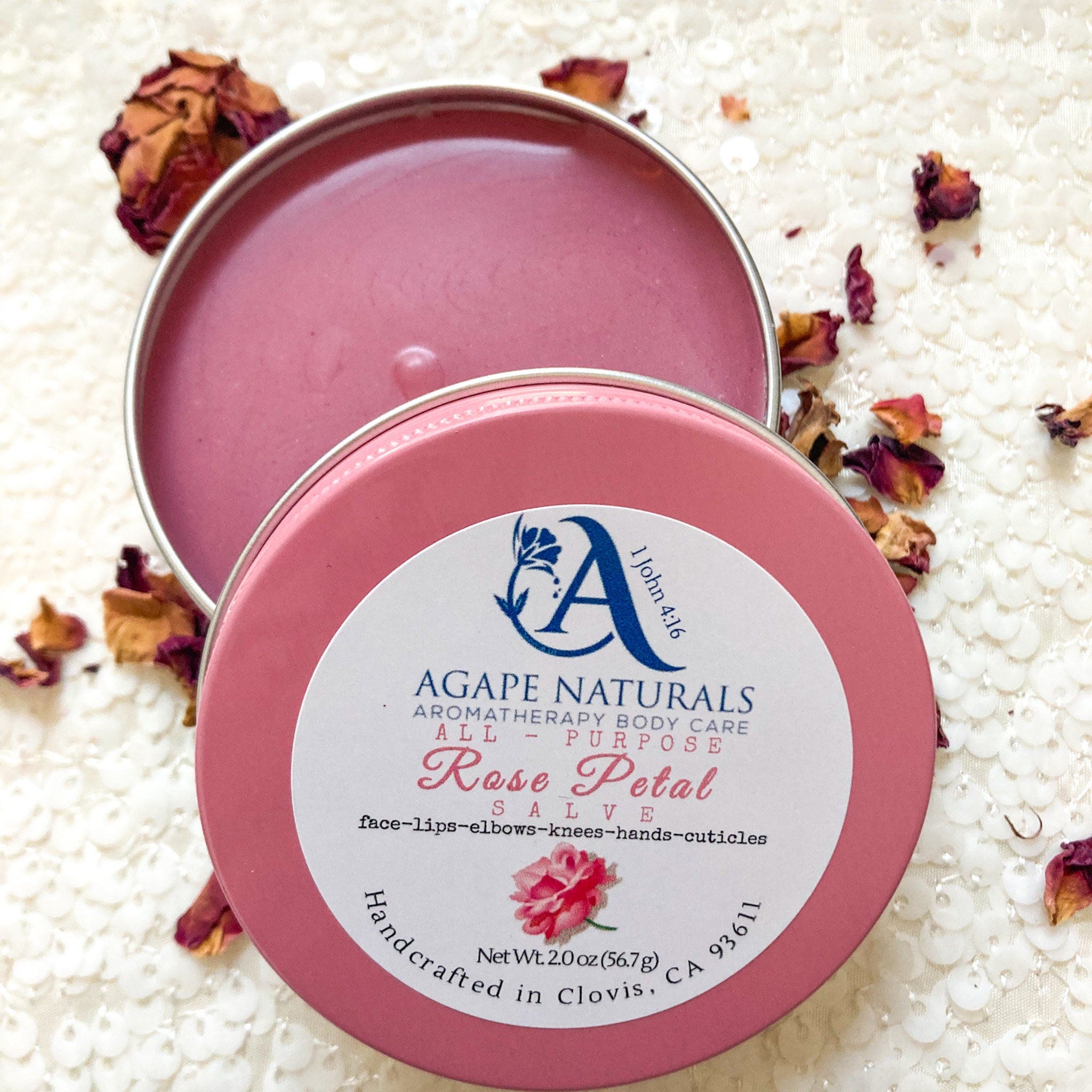 Body balm with pink salve, pink tin, and Agape Naturals label, surrounded by rose petals.