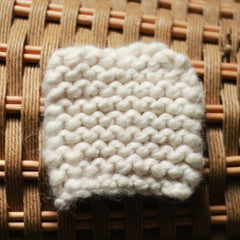 Knitted swatch of off-white yarn with visible stitch pattern resting on woven background