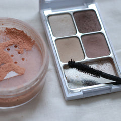 Eyeshadow palette with brown, beige, and tan shades, black mascara brush, and pink loose powder in a clear container