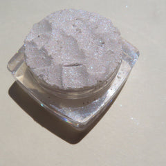 Side view of loose white eyeshadow with violet shimmer in a clear jar, showcasing sparkly glitter particles.