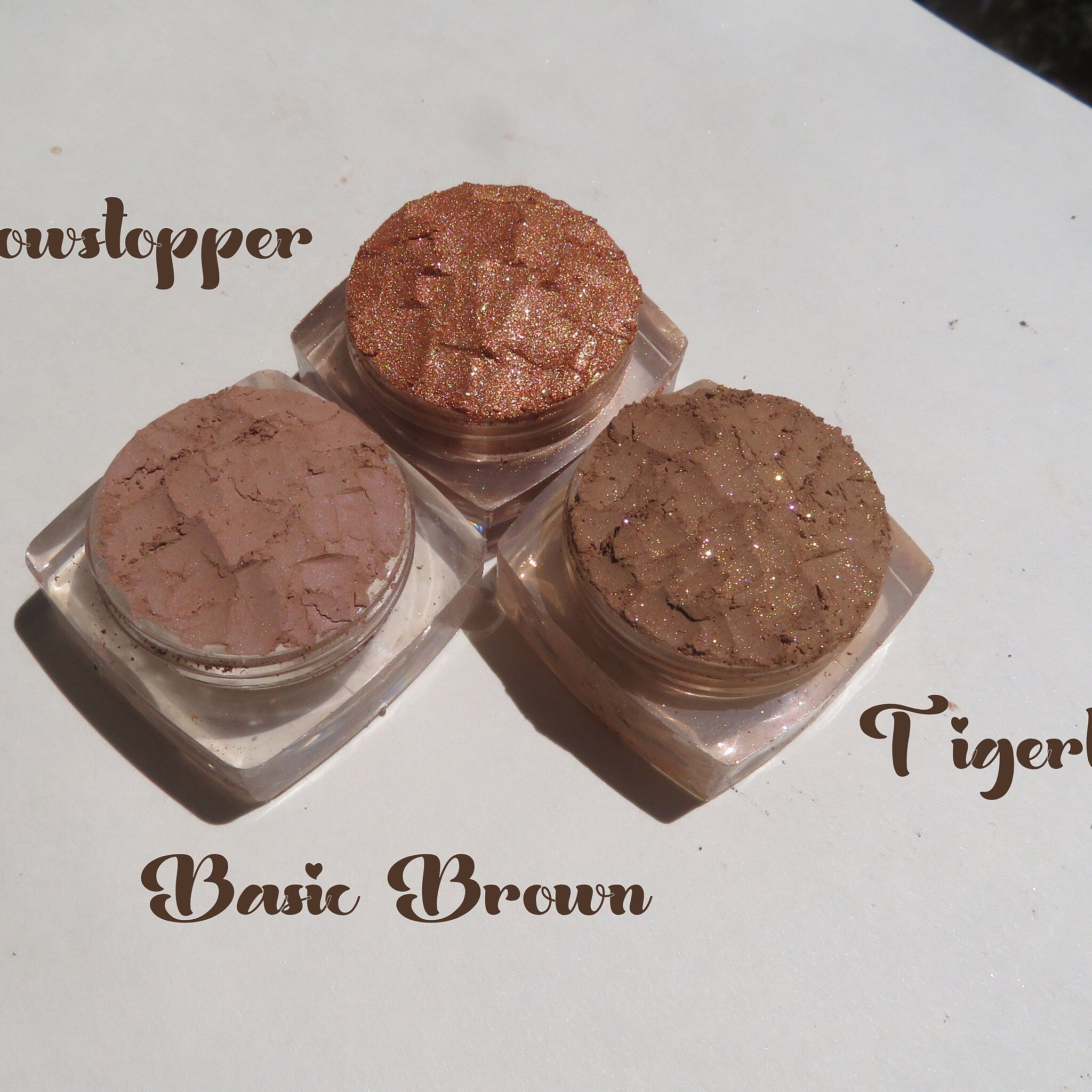 Three round mineral eyeshadow pans in fall shades of warm brown, bronze, and gold shimmer, arranged on white surface.