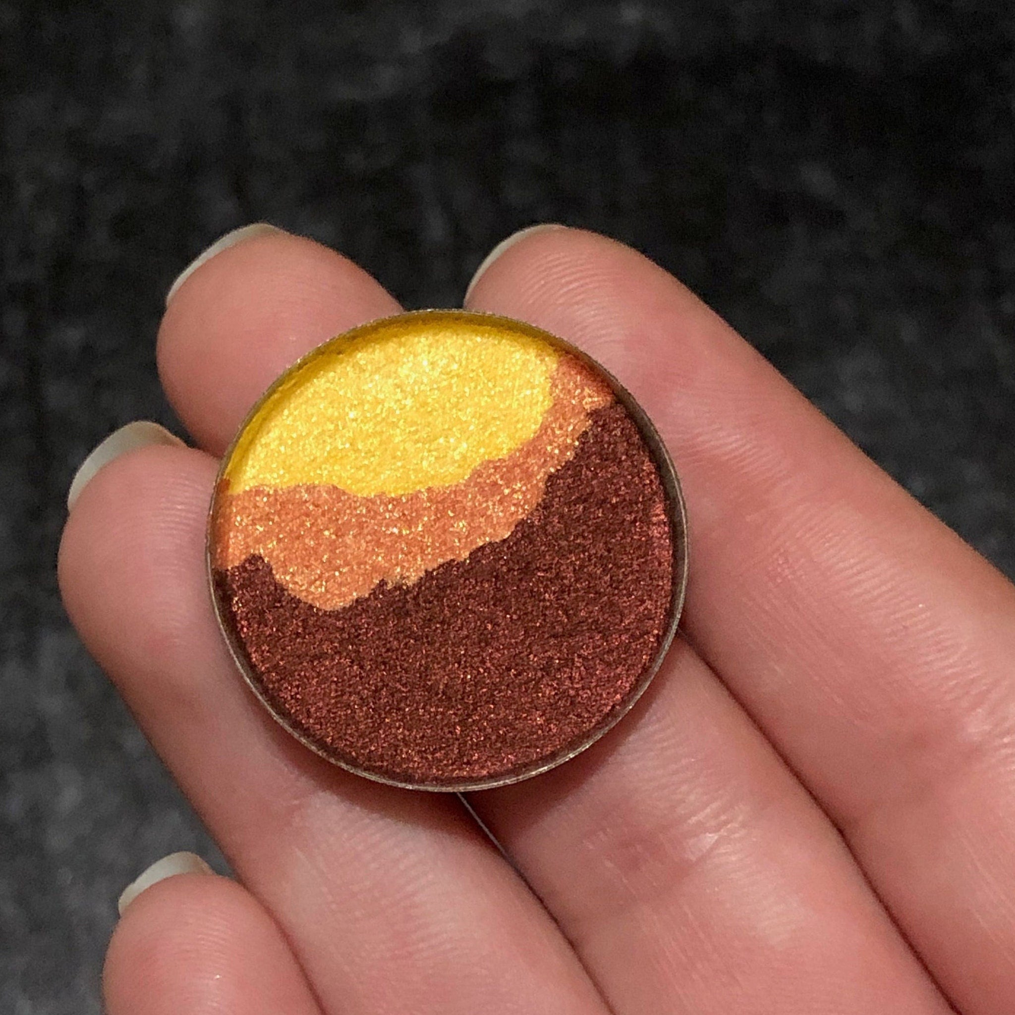 Eye makeup eyeshadow with gold, orange, and brown shades in a round compact held in fingers