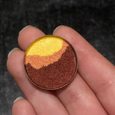 Eye makeup eyeshadow with gold, orange, and brown shades in a round compact held in fingers