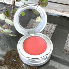 Blush compact with coral powder, silver metal case, and open lid on reflective surface