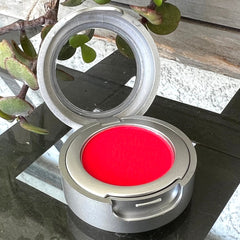 Makeup compact with red color, silver metal case, and open mirror lid on a patterned surface