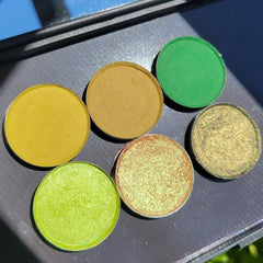 Side view of six green eyeshadow pans in earthy herb shades with smooth matte and shimmery finishes