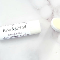 Lip balm tube, white with black text, "Rise & Grind" label, on a marble surface