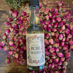 Rose water bottle with clear liquid and label, surrounded by pink rose buds and petals, Madame Phoenix's brand visible