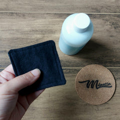 Reusable black cloth pad held by hand, with a bottle and cork coaster with "Mènessa" text on wood surface