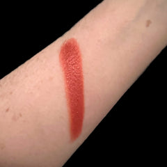 Lipstick swatch, reddish-orange color, textured finish, applied on arm