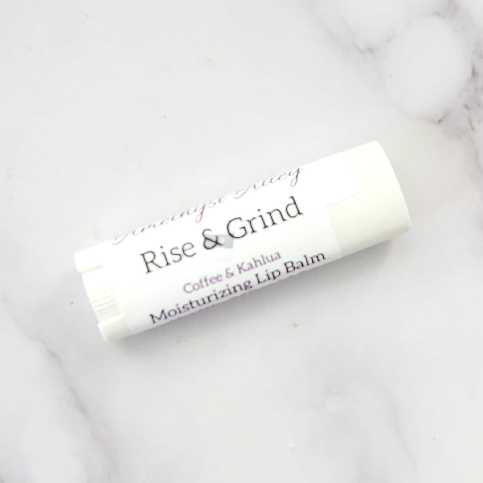 Lip balm tube with white and purple label, "Rise & Grind" text, on marble surface