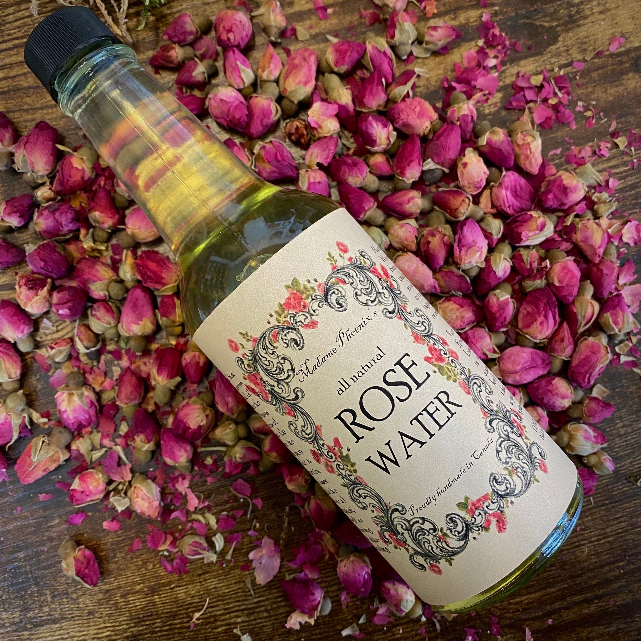 Rose water bottle with Madame Phoenix label, surrounded by pink rose buds on wood surface