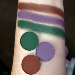 Eyeshadow swatches with green, purple, and brown colors applied on skin, showing circular pans