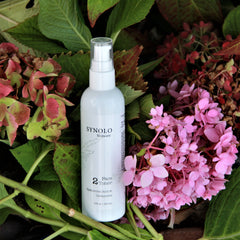 Face toner bottle, white plastic, Synolo Skincare label, surrounded by pink and green flowers