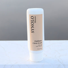 Citrus Scrub bottle, white plastic, beige label with Synolo Skincare branding, upright on marble surface