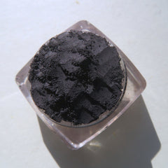 Black powder eyeshadow in clear plastic container, textured dark pigment, angled view