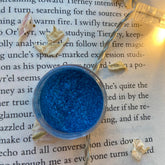 Eye makeup blue eyeshadow pot, glittery texture, clear plastic container on book page