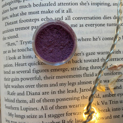 Eye shadow, dark purple powder in clear jar, on open book page, with fairy lights
