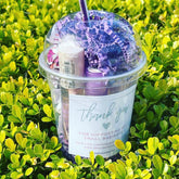 Lip gloss bundle in clear plastic cup with purple filler and DKWcosmetics label, on green foliage