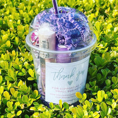 Lip gloss bundle in clear plastic cup with purple filler and DKWcosmetics label, on green foliage
