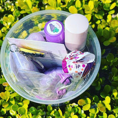 Purple lip gloss and keychain in clear plastic container, surrounded by green leaves