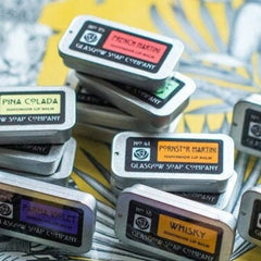 Lip balms in silver tins with colorful labels, Glasgow Soap Company branding visible, stacked together