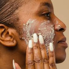 Facial scrub application, light beige paste, dark skin, white manicured nails, close up view