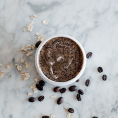 Coffee scrub in white bowl with brown granules and scattered coffee beans and oats