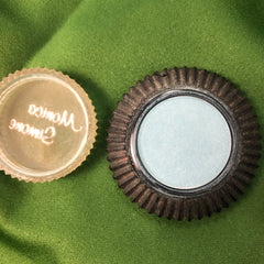 Eye shadow compact with blue powder, black rim, and clear lid with text on green fabric background