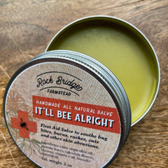 Salve tin with metal rim, green balm, and Rock Bridge Farmstead label on wood surface