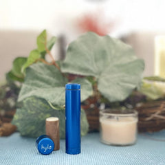 Blue cylindrical object, with "hyla" text on cap, set on blue textured surface with foliage