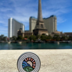 Round metal tin with white label, colorful logo, and "From Scratch Essentials" text, outdoors with blurred background