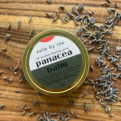 Balm tin with gold rim, green and white label, and lavender buds on wood surface sola by Isa balm