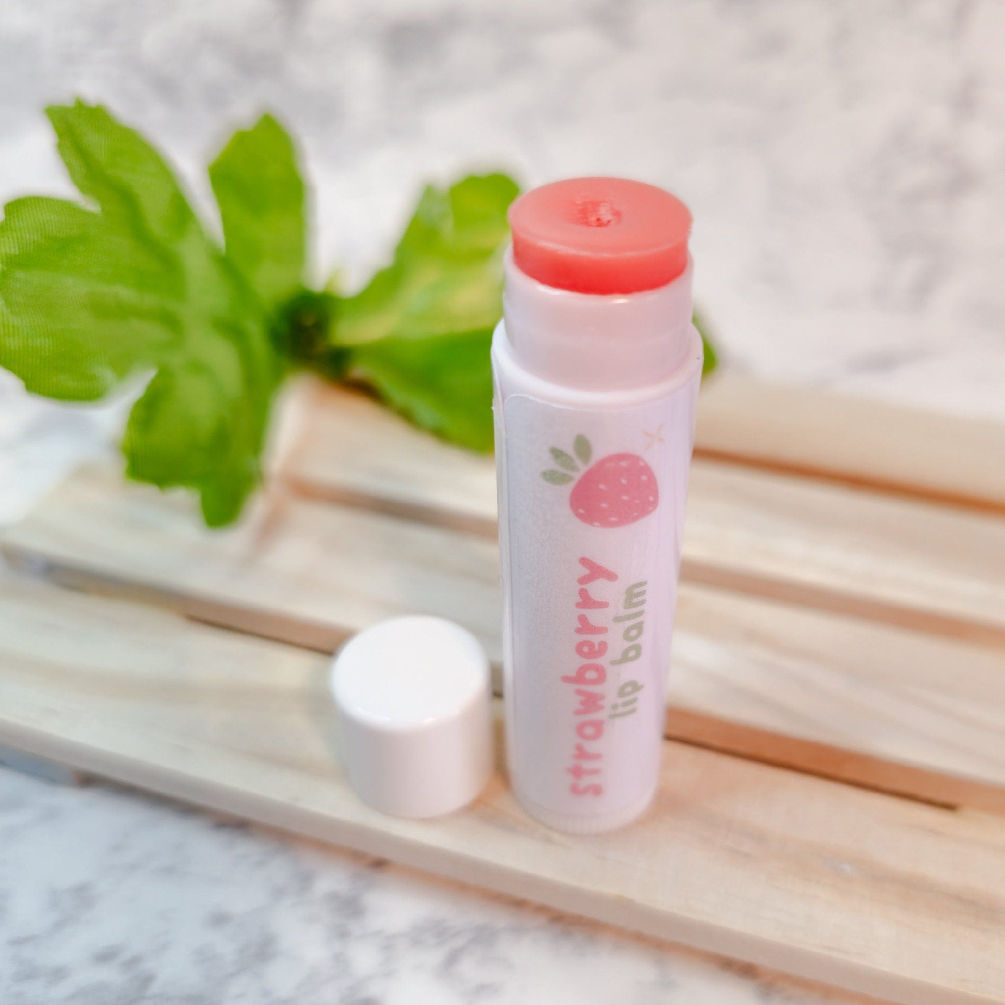 Lip balm tube with pink and white label, strawberry graphic, and open cap on wood
