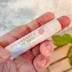 Strawberry lip balm in white tube with pink text and strawberry graphic, held in hand