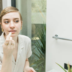 Lip balm application with white tube, fair skin, and gold jewelry in bathroom setting
