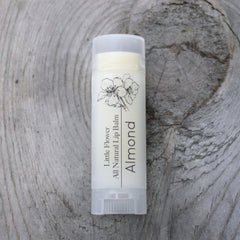 Lip balm tube with white label, floral graphic, and "Almond" text on wood surface