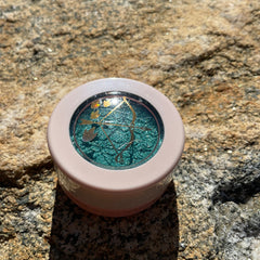 Eyeshadow compact with teal shimmer, gold bow and arrow design, and pink casing on rock surface