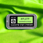 Lip balm tin with green text, silver metal case, and Glasgow Soap Company branding on green fabric
