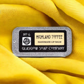 Lip balm tin with grey metal, black and cream text, on yellow fabric background Glasgow Soap Company