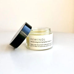 Eye cream jar with black lid, clear glass, and white label with REWINDI text