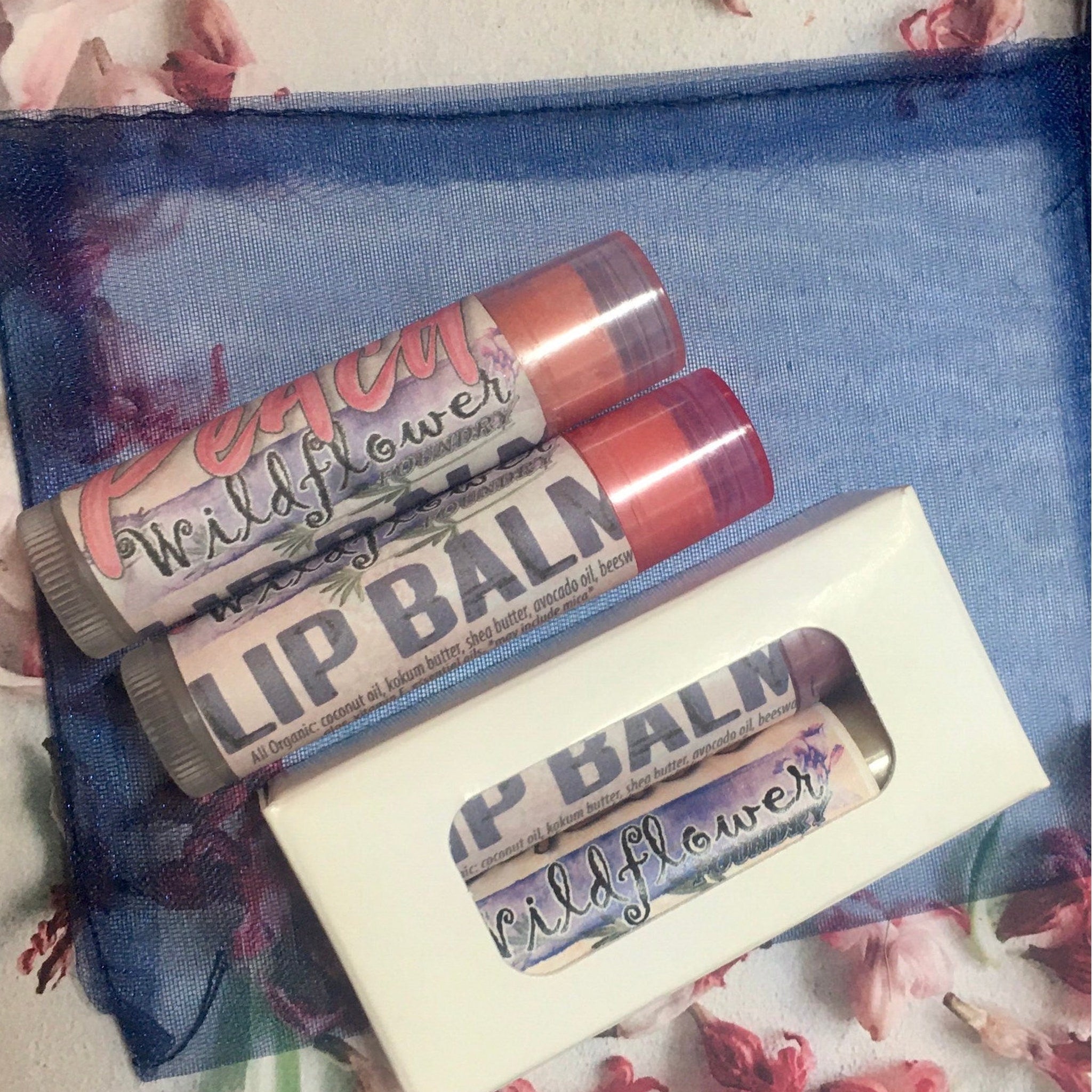 Lip balm tubes with pink and purple labels, white box, on blue mesh fabric