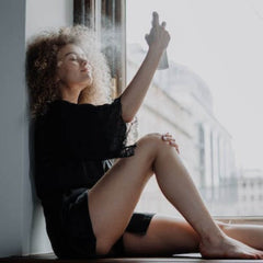 Woman spraying face with mist, black clothing, sitting by window, curly hair
