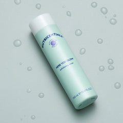 Exfoliating toner bottle, light green and white, with "Nutricentials" and "Here You Glow" text, with water droplets around it